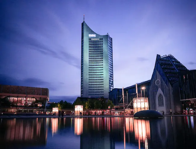 Blaue Abendstimmung am zentralen Teich in Leipzig, im Hintergrund spiegeln sich ein beleuchtetes Hochhaus und das Universitätsgebäude im Wasser.