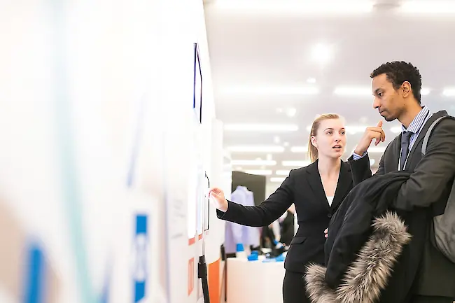 Eine Frau erklärt auf einer Messe einem Mann etwas auf einem großen Bildschirm an einem Messestand. Eine Frau erklärt auf einer Messe einem Mann etwas auf einem großen Bildschirm an einem Messestand.