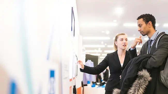Eine Frau erklärt auf einer Messe einem Mann etwas auf einem großen Bildschirm an einem Messestand.
