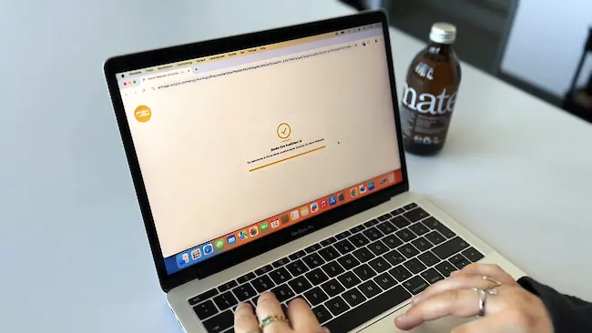 Ein Laptop zeigt auf dem Bildschirm eine Bestätigungsmeldung, während Hände die Tastatur bedienen; im Hintergrund steht eine Getränkeflasche.