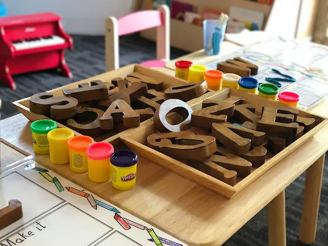 Ein hölzerner Aktivitätstisch in einem Klassenraum mit Schalen großer Holzbuchstaben und bunten Knete‑Töpfen, bereit für kreatives Kinderspiel.