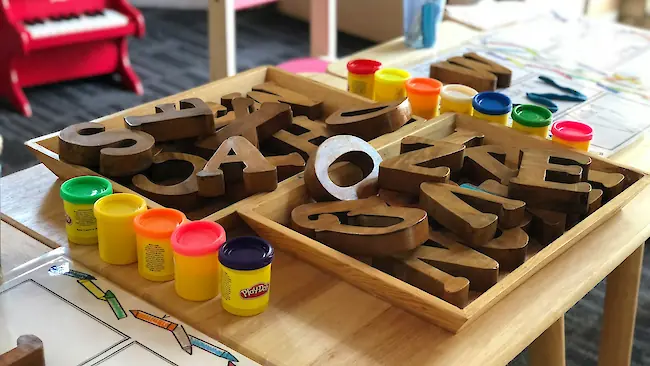 Ein hölzerner Aktivitätstisch in einem Klassenraum mit Schalen großer Holzbuchstaben und bunten Knete‑Töpfen, bereit für kreatives Kinderspiel.