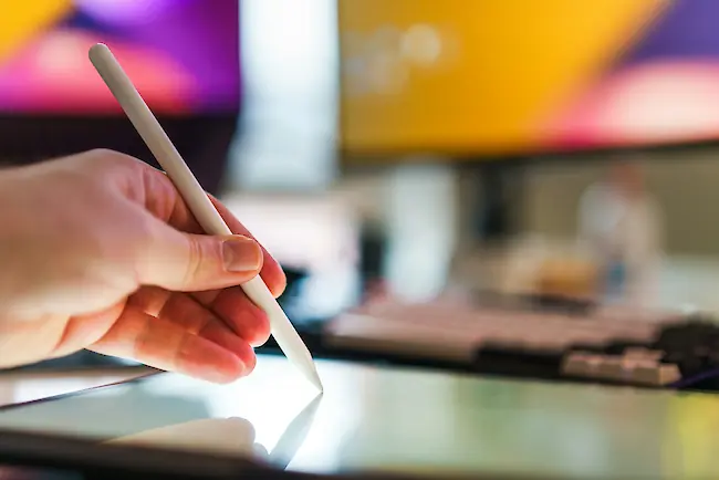 Eine Hand hält einen weißen Stylus und arbeitet am Bildschirm eines Grafiktabletts in einem unscharfen Arbeitsbereich mit Computerbildschirmen im Hintergrund. Eine Hand hält einen weißen Stylus und arbeitet am Bildschirm eines Grafiktabletts in einem unscharfen Arbeitsbereich mit Computerbildschirmen im Hintergrund.