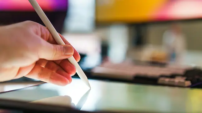 Eine Hand hält einen weißen Stylus und arbeitet am Bildschirm eines Grafiktabletts in einem unscharfen Arbeitsbereich mit Computerbildschirmen im Hintergrund.