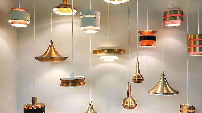 Eine Sammlung moderner Pendelleuchten in gebürstetem Messing und Weiß, in verschiedenen Höhen hängend vor einer neutralen Wand.